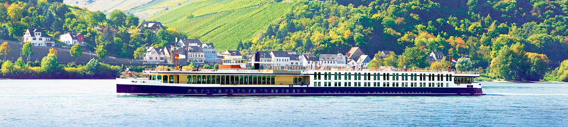 Exterior of a river cruise ship on the Rhine River with a castle on a green hill in the background.
